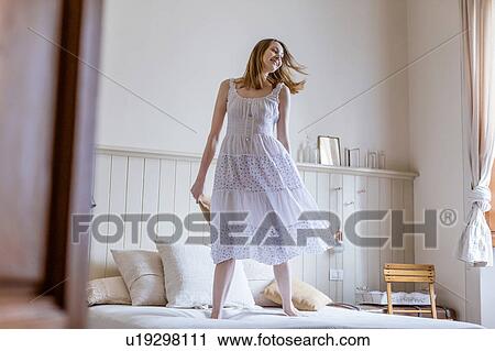 Stock Image - Woman wearing white dress jumping on bed. Fotosearch