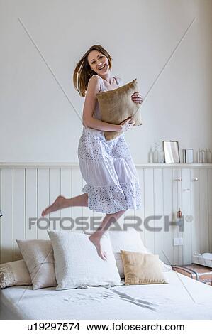 Picture - Woman wearing white dress jumping on bed looking at camera smiling. Fotosearch