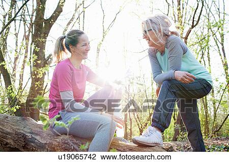 Stock Image - Women in forest sitting on fallen tree face to face smiling. Fotosearch