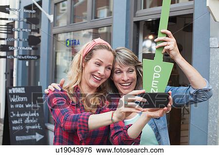 Women in front of shop holding open sign taking selfie with smartphone View Large Photo Image Stock Photograph - Women in front of shop holding open sign taking selfie with smartphone. Fotosearch