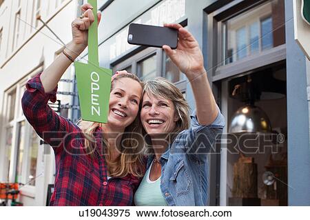 Stock Photography - Women in front of shop holding open sign taking selfie with smartphone. Fotosearch