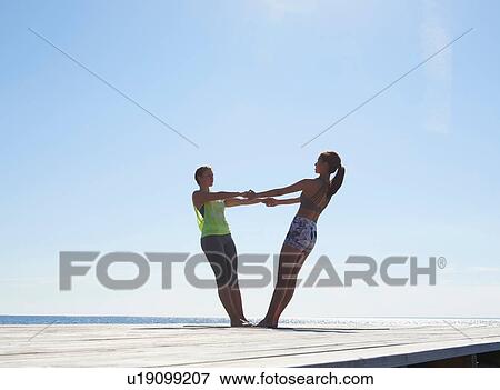 Stock Photo - Women on pier face to face holding hands leaning back exercising. Fotosearch