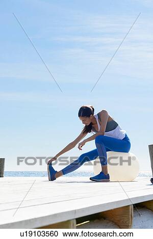 Stock Image - Women wearing sports clothes sitting on pier on exercise ball stretching foot. Fotosearch