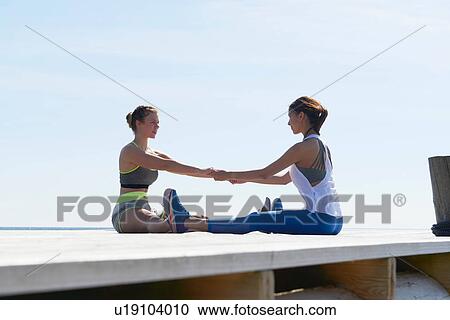 Stock Image - Women wearing sports clothes sitting on pier face to face holding hands exercising. Fotosearch