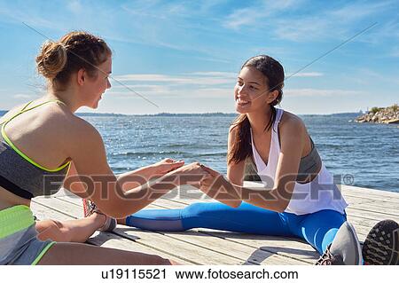 Stock Image - Women wearing sports clothes sitting on pier holding hands face to face smiling. Fotosearch