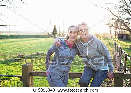 Stock Image - Women wearing sports clothing leaning against fence looking at camera hugging and smiling. Fotosearch