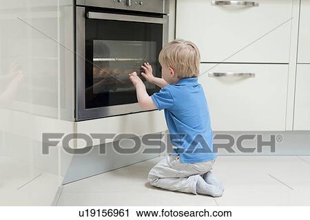 Stock Image - Young boy sitting in front of oven, looking through glass, rear view. Fotosearch