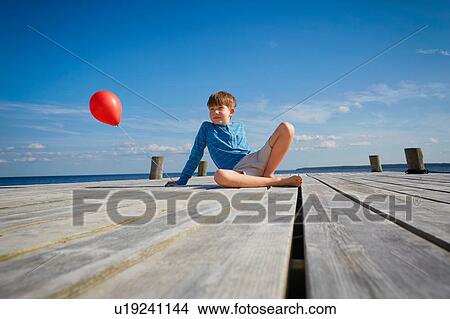 Picture - Young boy sitting on wooden pier, holding red helium balloon. Fotosearch