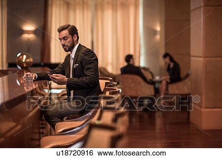 Stock Photograph - Young businessman sitting at hotel bar reading smartphone texts. Fotosearch