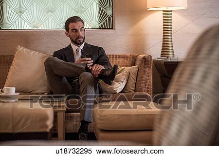 Young businessman sitting on hotel lobby sofa using smartphone View Large Photo Image Stock Photography - Young businessman sitting on hotel lobby sofa using smartphone. Fotosearch