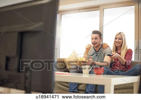 Stock Image - Young couple at home, sitting on sofa, watching television, excited. Fotosearch