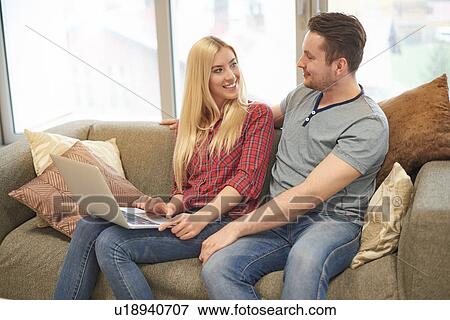 Young couple at home, sitting on sofa, using laptop View Large Photo Image Stock Photo - Young couple at home, sitting on sofa, using laptop. Fotosearch