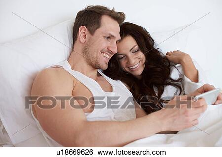 Stock Photograph - Young couple sitting up in bed browsing digital tablet. Fotosearch