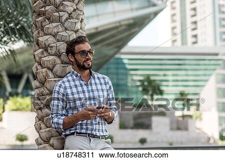 Stock Image - Young man leaning against palm tree using smartphone, Dubai, United Arab Emirates. Fotosearch