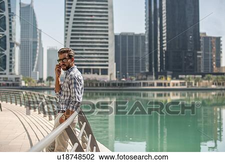 Stock Image - Young man leaning against waterfront railings talking on smartphone, Dubai, United Arab Emirates. Fotosearch