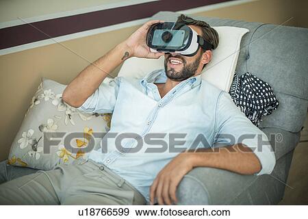 Stock Photo - Young man on armchair looking through virtual reality headset. Fotosearch
