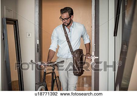 Stock Image - Young man pushing bicycle through apartment door. Fotosearch