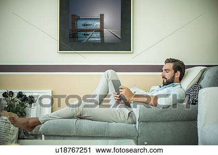 Stock Image - Young man reclining on hotel room chaise longue using digital tablet, Dubai, United Arab Emirates. Fotosearch