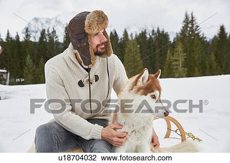 Young man wearing trapper hat petting husky in snow, Elmau, Bavaria, Germany View Large Photo Image Stock Image - Young man wearing trapper hat petting husky in snow, Elmau, Bavaria, Germany. Fotosearch