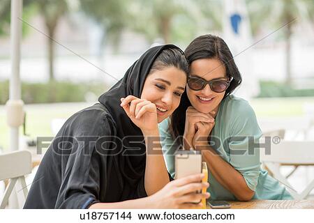Picture - Young middle eastern woman wearing traditional clothing reading smartphone text with female friend at cafe, Dubai, United Arab Emirates. Fotosearch