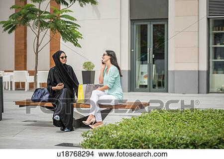 Young middle eastern woman wearing traditional clothing sitting on bench with female friend, Dubai, United Arab Emirates View Large Photo Image Picture - Young middle eastern woman wearing traditional clothing sitting on bench with female friend, Dubai, United Arab Emirates. Fotosearch
