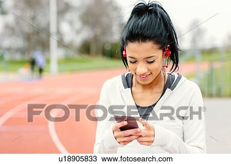 Stock Image - Young woman beside running track, holding smartphone, wearing earphones, smiling. Fotosearch