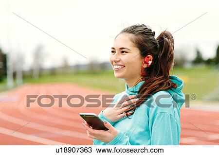 Stock Image - Young woman beside running track, holding smartphone, wearing earphones, smiling. Fotosearch