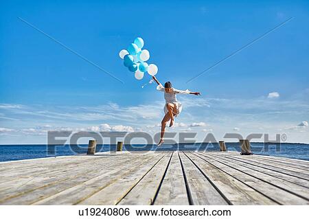 Stock Photograph - Young woman dancing on wooden pier, holding bunch of balloons. Fotosearch