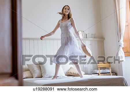 Stock Image - Young woman in long white dress jumping on bed looking at camera smiling. Fotosearch