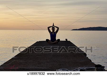 Stock Image - Young woman sitting on pier, in yoga position, at dusk. Fotosearch