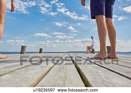 Stock Photo - Young woman sitting on post on wooden pier, looking at friends standing further away. Fotosearch