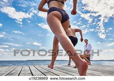 Young woman sitting on post on wooden pier, watching friends running towards edge, low angle view View Large Photo Image Stock Photo - Young woman sitting on post on wooden pier, watching friends running towards edge, low angle view. Fotosearch