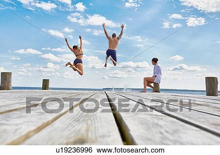 Stock Photograph - Young woman sitting on post on wooden pier, watching friends as they jump into sea. Fotosearch