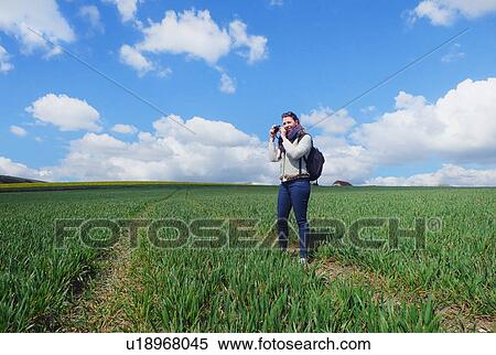 Stock Photography - Young woman standing in field, taking photograph of landscape. Fotosearch