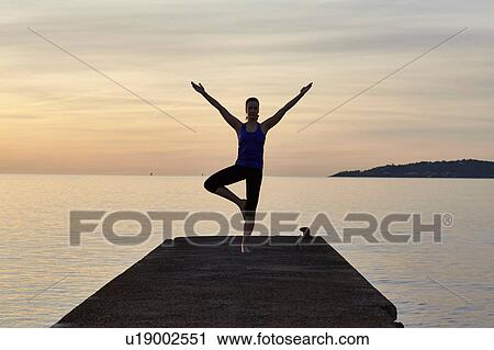 Young woman standing on pier, in yoga position, at sunset View Large Photo Image Stock Image - Young woman standing on pier, in yoga position, at sunset. Fotosearch