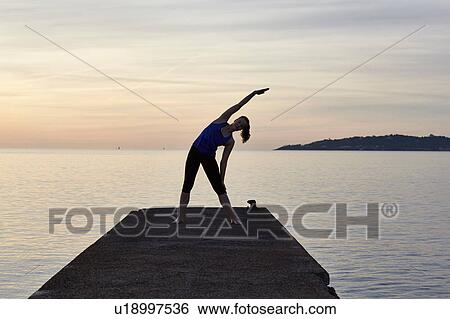Stock Photograph - Young woman standing on pier, in yoga position, at sunset. Fotosearch