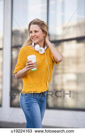 Stock Image - Young woman walking outdoors, holding takeaway coffee cup, wearing headphones around neck, smiling. Fotosearch