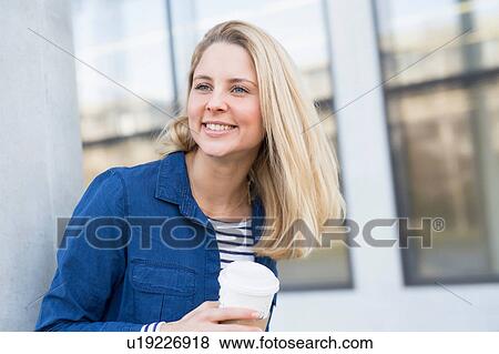 Stock Photo - Young woman walking outdoors, holding takeaway coffee cup, smiling. Fotosearch