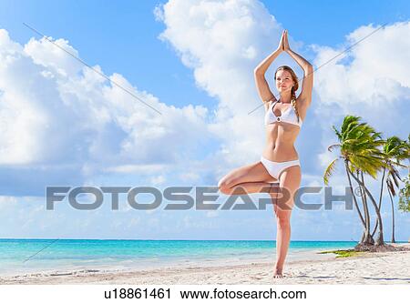 Stock Image - Young woman wearing bikini practising yoga tree pose on beach, Dominican Republic, The Caribbean. Fotosearch