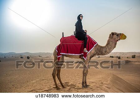 Stock Image - Young woman wearing traditional middle eastern clothes riding camel in desert, Dubai, United Arab Emirates. Fotosearch