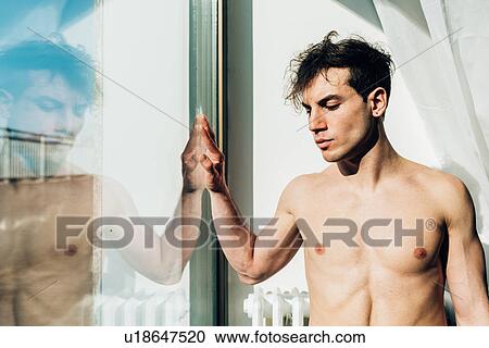 Stock Image - Bare chested man looking out of window. Fotosearch