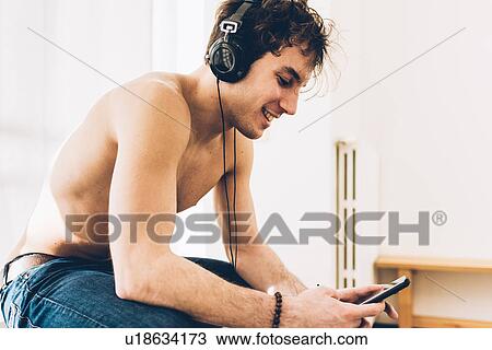 Stock Image - Bare chested man wearing headphones looking down at smartphone smiling. Fotosearch