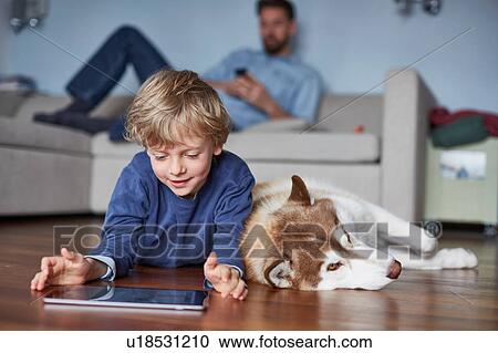 Stock Image - Boy lying on living room floor with husky using digital tablet. Fotosearch