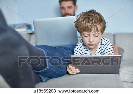 Stock Image - Boy lying on sofa with father using technology. Fotosearch