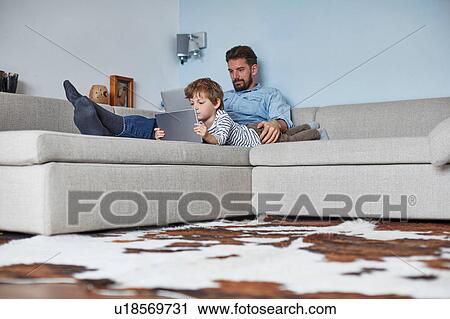 Stock Image - Boy lying on sofa with father using technology. Fotosearch