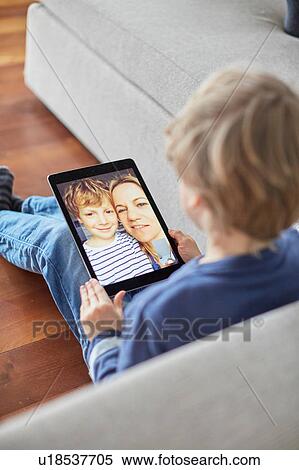 Stock Photography - Boy sitting on floor talking to mother and brother on digital tablet video call. Fotosearch