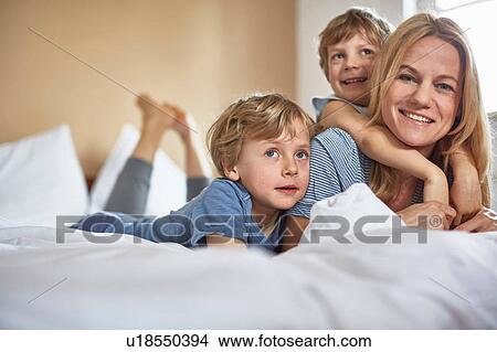 Picture - Boys on bed lying on top of mother looking at camera smiling. Fotosearch