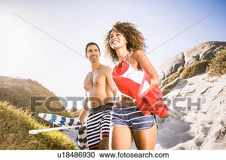 Stock Image - Couple on sand dune carrying beach umbrella and towel, Cape Town, South Africa. Fotosearch