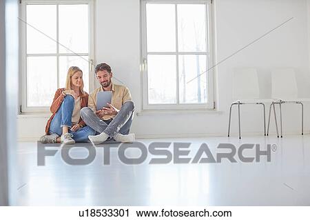 Stock Image - Couple sitting on floor browsing digital tablet in empty new home. Fotosearch
