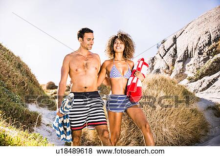 Stock Photo - Couple walking down sand dune carrying beach towel and umbrella, Cape Town, South Africa. Fotosearch